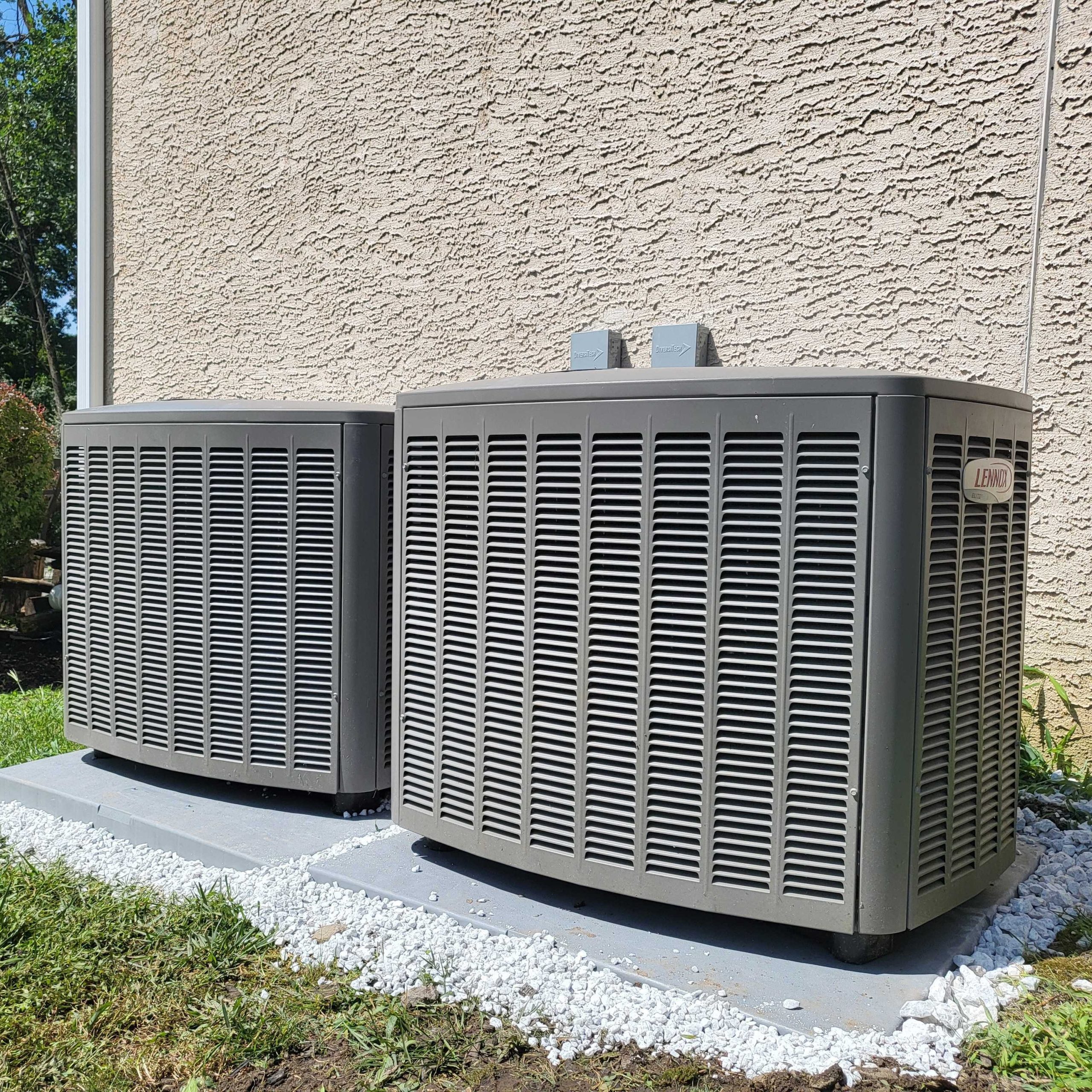 Two grey Lennox AC units sit on a concrete pad with white gravel in a yard.
